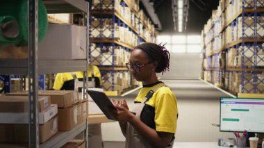 Tabletle ve dağıtım merkeziyle birlikte sipariş işleme görevlerinde çalışan siyahi kadın sanayi katibi verimli bir depolama operasyonu sağlıyor. E-ticaret dağıtımı için veri paketlerini yönetiyor. Kamera A.