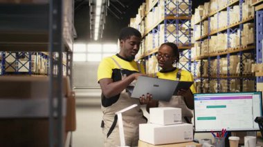 Lojistik merkezindeki sipariş işlemleri için dizüstü bilgisayardaki paket verileriyle ilgilenen siyah takım üyeleri, dağıtım protokolleri için palet taşıma etiketlerini doğruladılar. Verimli bir e-ticaret deposu. Kamera A.