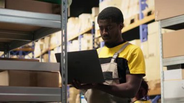 Genç zenci çalışanlar büyük bir depo tesisinde raflarda paketleri ayırıyorlar. Envanteri paketlenmiş malların etrafındaki bilgisayar ile yönetiyorum ve yerine getirmek için dağıtıma hazırım. Kamera B.