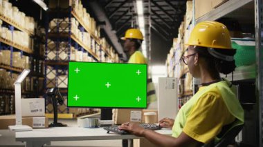 Güvenlik yeleği giymiş bir kadın bilgisayardaki yeşil ekranın yanındaki barkodları kontrol ediyor, e-ticaret dağıtım operasyonları üzerinde çalışıyor ve kask takıyor. Büyük bir depo rafı. Kamera A.