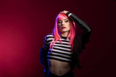 Çizgili bluzlu şık bir kadın moda stüdyosunda güçlü poz veriyor. Kolunu kaldırıp dik dik bakıyor. Renkli saç stili ve deri ceketli kendine güvenen bir model..