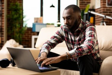 Ciddi bir erkek yönetici rahat bir ofiste oturmuş dizüstü bilgisayarda yazı yazıyor ve mali verileri inceliyor. Afrikalı Amerikalı girişimci üretken kalıyor, şirket büyümesini sürdürmek için uzaktan çalışıyor..