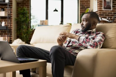 Düşünceli iş adamının portresi modern oturma odasındaki koltukta dizüstü bilgisayarı ile dinleniyor. Afrika kökenli Amerikalı girişimci sadece hayata, duygulara ve gelecekteki kararlara bakıyor..