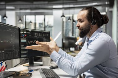 Borsacı, borsadaki müşterilerle mikrofonla konuşurken alım satım siparişlerini yürütüyor. Müşterilere finansal hedeflere ulaşmalarında yardım eden, yatırım fırsatları konusunda danışmanlık yapan adam