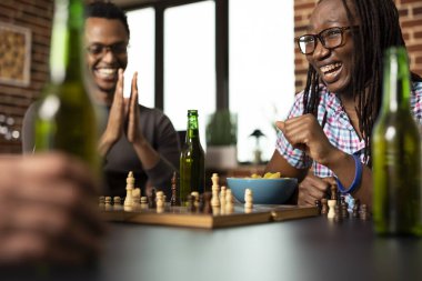 Genç Afrikalı Amerikalı kadın satranç zaferini sevinçle kutlarken erkek arkadaşı onun için alkış ve tezahürat yapıyor. Kapalı alan ortamında yakın arkadaşlar arasında keyifli masa oyunu oturumları.