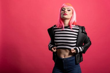 Pembe peruklu ve çizgili bluzlu şık beyaz kadın izole edilmiş arka planda kameraya bakıyor. Deri ceketli kendine güvenen kadın model, stüdyoda moda fotoshoot sırasında cesur bir tavır sergiliyor..