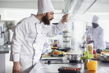 Üniformalı profesyonel bir aşçı sobanın üstündeki tavadan lezzetli bir sos tadıyor ve lezzetlerin mükemmel dengelenmesini sağlıyor. Modern bir restoranın mutfak tezgahında lezzetli yemekler kaynıyor..