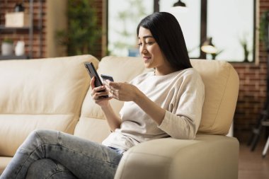 Modern apartmandaki koltukta oturan neşeli bayan girişimci akıllı telefon ve banka kartını dijital ödeme için kullanıyor. Cep telefonundan online alım yapan Asyalı iş kadını.