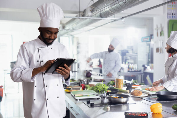 Doing culinary research, african american chef in a restaurant kitchen uses tablet to find new dish tips while preparing fresh ingredients. Man cooking gourmet meal, delicious raw materials.