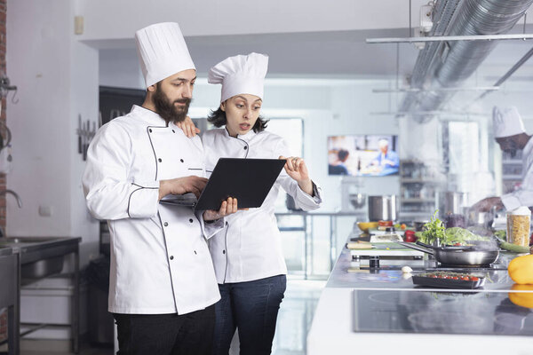 Professional chefs researching recipes on laptop and preparing gourmet dishes in modern restaurant kitchen. Fresh ingredients, kitchenware and a stove surround the busy culinary environment.