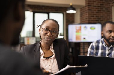 Elinde not panosu olan siyah bir bayan yönetici. Başlangıç ofisindeki çalışanların performans ölçümlerini inceliyor. Afrikalı Amerikalı iş kadını grup toplantısı sırasında iş arkadaşlarıyla tartışıyor..