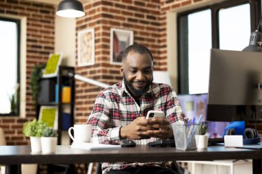 Modern ofis masasında oturan ve akıllı telefon kullanan mutlu bir Afrikalı Amerikalı 'nın portresi. Serbest çalışan siyahi adam işten dinleniyor, mobil cihaz kullanarak çevrim içi ağ kuruyor.
