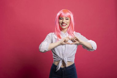 Gündelik giysiler içinde mutlu pembe saçlı bir kadın elleriyle sevgi ve neşe ifade eden bir kalp şekli oluşturur. Güzel genç bayan pembe arka planda romantik bir tebessümle yalnız duruyor..