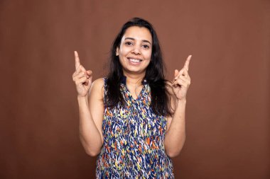 Kahverengi arka planda duran neşeli kadın, gülümseyerek ve işaret parmaklarıyla yükselerek terfi için boş alana doğru hareket ediyor. Kafasını kaldırmış neşeli bir kadın.