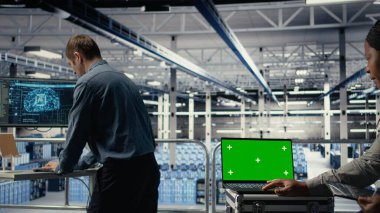 Veri merkezi teknisyeni, yeşil ekran laptopuyla derin öğrenme sürecini gözlemler ve operasyonel ölçümleri gözden geçirir. Denetleme sırasında nöral ağların raporlarını gözden geçirmek için A kamerasını kullanan yönetici