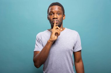 Siyahi bir adam mavi arka planda duruyor, sessiz bir jest yaparak işaret parmağıyla dudaklara bastırıyor, gizliliği işaret ediyor. Afro-Amerikan erkek kişi sessizlik talep ediyor, sağduyulu olmayı öneriyor..