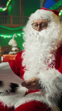 Dikey video. Mutlu adam, Noel karakterini canlandırıyor. Çocuklardan mektuplar okuyor, şapka ayarlıyor. Sahte sakal takmış, Noel Baba gibi gülümseyen, çocukların dilek listelerinin keyfini çıkaran, kamera A