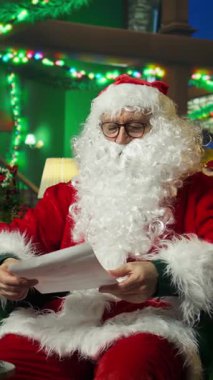 Dikey video Noel Baba çocuklardan gelen mektuplara bakar, onları selamlar ve karşılar. Noel Baba kostümlü gülümseyen kişi çocukların Noel için yazdığı dilek listesini okur, el sallar, selamlar.