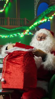 Noel Baba 'nın Noel çantasını hediyelerle doldurduğu dikey video. Sahte sakallı yaşlı bir adam hediye paketliyor, seyahat etmeye ve çocukları ziyarete hazırlanıyor.