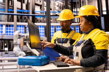 Mühendislik ekibi otomatikleştirilmiş fabrika operasyonlarını denetliyor, dijital kontrolleri kullanarak robotik silahlar ve endüstriyel tesis verimliliğini arttırıyor. Siyah kadın ve erkek akıllı üretim tesisinde işbirliği yapıyor..