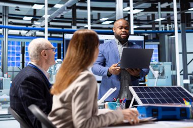 Güneş panellerindeki Afrikalı Amerikalı işadamı yatırım stratejisini değerlendirmek için not defterindeki verilere bakıyor. Fotovoltaik fabrikasında imalat genişletme seçeneklerini gözden geçirmek için dizüstü bilgisayar kullanan yatırımcı