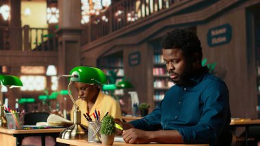 Afrikalı Amerikalı öğrenci kütüphanede anahtar sözcüklerin ve cümlelerin altını çizerek üniversitenin kampüs çalışma alanında fosforlu kalemle bir özet hazırlıyor. Sınavlar için akademik araştırma. Kamera B.