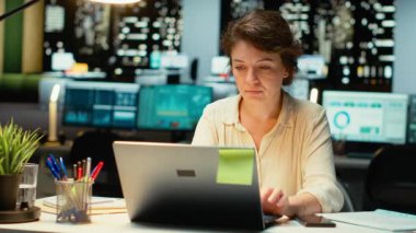 Kadın çalışan telefonunu kontrol edip laptopunu kapatıyor, fazla mesai yaptıktan sonra gece geç saatlerde ofisten çıkmaya hazırlanıyor. Yoğun iş yoğunluğu mesai sonrası kalmasını sağlıyor. Kamera B.