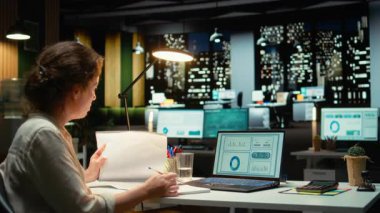 Yönetici, bir sonraki toplantı, büyüme planı için ikna edici bir sunum yaratmak amacıyla strateji odasında bir rapor hazırlamak ve güncellemeleri gece vakti belgelemek için rakamları araştırıyor. Kamera A.