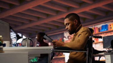 Tekerlekli sandalyedeki yönetici, uygulamayı geliştirmek için yenilikçi başlangıç ofisinde tablet kullanıyor. Belden aşağısı felçli özel yazılım geliştirme ajansı çalışanı cihaz üzerindeki kodu kontrol ediyor, kamera B