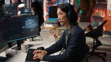 Yapay Zeka 'nın başlangıç ofisindeki kıdemli programcı kulaklık takıyor, otomasyonu geliştirmek için makine öğrenme modellerini inceliyor. Çalışırken müzik dinleyen Asyalı CTO yöneticisi, kamera A