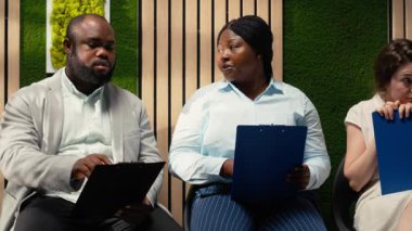 Röportaj öncesinde koridorda oturan kendine güvenen iş başvuruları, özgeçmişlerini gözden geçirip yetenek edinme toplantısı sırasında HR çalışanlarıyla birlikte iş sorularına hazırlanıyorlar. Kamera B.