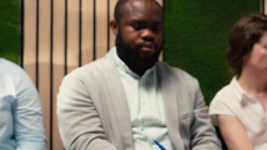 Koridorda evraklar ve özgeçmişlerle bekleyen üç iş arayan insan kaynakları ekibiyle resmi bir görüşme için hazırlanıyor. Başvuranlar koridorda kuyrukta sabırsızlanıyorlar. Kamera B.