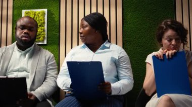 Röportaj öncesinde koridorda oturan kendine güvenen iş başvuruları, özgeçmişlerini gözden geçirip yetenek edinme toplantısı sırasında HR çalışanlarıyla birlikte iş sorularına hazırlanıyorlar. Kamera A.