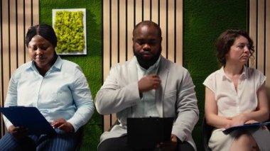 Koridorda CV dosyalarıyla bekleyen çok ırklı iş arayanlar, İK ile resmi bir röportaj için hazırlanıyorlar. Başvuranlar, kafa avcılığı sırasında sabırsızlık ve kendine güveni yansıtıyor. Kamera A.