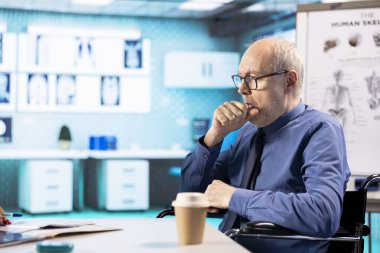 Solunum sorunuyla ilgilenen ve kabinede öksüren hasta tekerlekli sandalye kullanıcısı, muayene sırasında astım veya zatürreeden muzdarip. Sağlık görevlisi yaşlı adama enfeksiyon konusunda yardım ediyor..