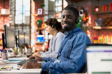 Müşterilere donanım sorunuyla ilgili teknik sorulara cevap vermek için kulaklık kullanan neşeli çağrı merkezi çalışanının portresi. Sorunları çözmek için bilgisayar kullanarak müşteri destekleme işi yapan iyimser bir adam