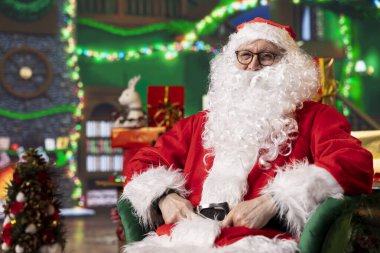 Çocukların merakını gidermek için Noel Baba karakterini oynayan gülümseyen bir şovmen portresi. Noel Baba gibi giyinmiş neşeli bir adam Noel süsleri odasında mevsimlik hediye teslimatına hazırlanıyor.