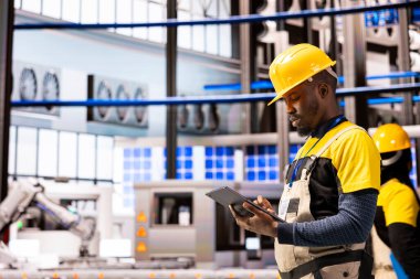 Tabletli siyah erkek mühendis akıllı üretim tesisinde üretim hattı makinelerinin bakımını yapıyor. Otomatik fabrikada robotik kol ve taşıma bantlarını kontrol eden Afrikalı Amerikalı teknisyen