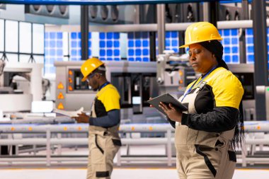 Afrikalı Amerikalı teknisyenler sanayi 4.0 fabrikasını denetliyor ve üretim sürecini değerlendiriyor. Kadın mühendis tablet tutuyor, akıllı fabrika değerlendirmesi ve kalite kontrolü yapıyor..
