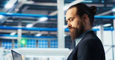 Fotovoltaik fabrikası çalışanı verileri analiz ediyor, sistem sağlık ölçümlerini dizüstü bilgisayarla inceliyor. Güneş panelleri fabrika çalışanlarının teknik sorunlarını not defteri ve kamera B kullanarak çözüyor.