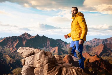 Doğa aşığı nefes kesen bir dağ sırasının kenarında duruyor, yükselen zirvelerin ve uçsuz bucaksız gökyüzünün manzaralı arka planına bakıyor. Genç adam macera ve açık hava keşiflerini seviyor..