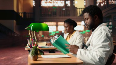 Siyahi tıp öğrencileri kütüphanede bilgi ve sınıf notlarını karşılaştırıyor, birlikte çalışarak ihtisas yapıyorlar. Öğrenciler ameliyat notlarını okuyarak sağlık yöntemlerini öğreniyorlar. Kamera A.