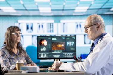 Muayenedeki hasta ve tıp uzmanı, yeni tedavi planı için tedavi ve klinik ilaçlar hakkında görüştü. Erkek doktor ilaçlara işaret ediyor ve günlük dozu açıklıyor..