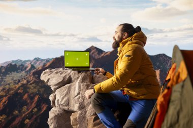 Dağcı memnuniyetle zirvede durur ve taklit ekran kullanır. Yürüyüşçü dağın tepesinde canlı havayla nefes kesici manzaraya hayran kalır. Manzara çevresi kararlılığı vurguluyor.