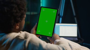 Afrikalı Amerikalı genç yetişkin, o koltukta dinlenirken oturma odasında aletlerini kullanarak yeşil ekrana bakıyor. Erkek kişi kendini rahat ve uzak işten memnun hisseder. Kamera A.