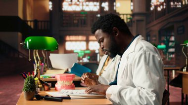 Afrika kökenli Amerikalı dişçi, insan dişlerinin plastik modelini inceliyor, kampüs kütüphanesinde stomatoloji çalışmaları yapıyor. Erkek öğrenci dişçiliği ya da oral sağlığı öğrenir. Kamera A.