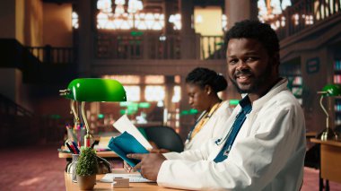 Afrikalı Amerikalı tıp öğrencisi kütüphanede yeşil lambaların altında anatomi materyalleri ve dava dosyaları hakkında araştırma yapıyor. Sınav hazırlıkları tıp fakültesindeki kararlılığın altını çiziyor. Kamera A.