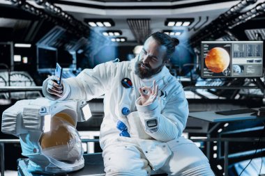 Astronot, arka planda Mars görev ekranı parlayan uzay istasyonunun içinde selfie çekiyor. Yıldız gemisi mürettebatı Kızıl Gezegen 'e inmeden önce cep telefonu kullanarak el hareketi yapıyor.