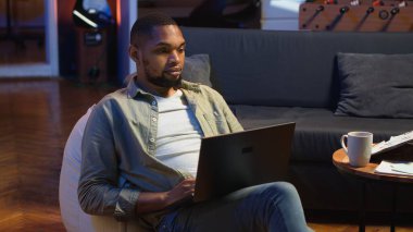 Genç siyahi biri dizüstü bilgisayarında uzaktan kumandalı bir video ile evde oturuyor, görevleri çözmek için online bir konferans web sitesine katılıyor. Oturma odasında telekonferans. Kamera B.