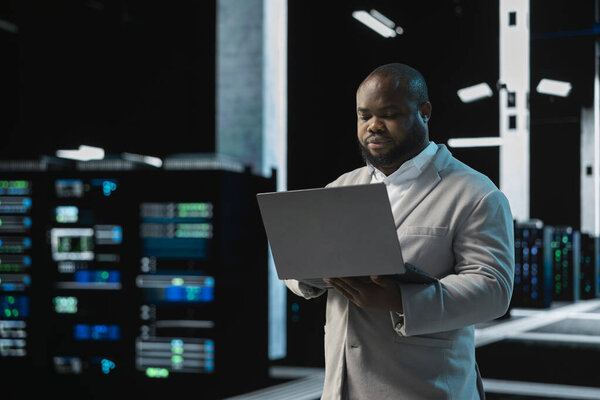 Software developer in data center using laptop, reading error logs, doing checkup to prolong hardware life span. Server hub computer scientist monitoring infrastructure rigs using notebook